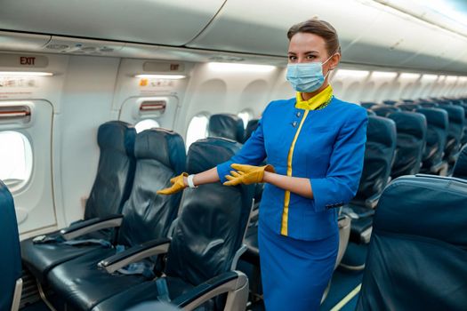 Woman flight attendant wearing protective medical mask and air hostess uniform while gesturing towards passenger chair in airplane
