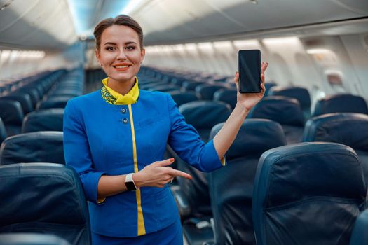 Joyful stewardess with smartphone standing in airplane cabin