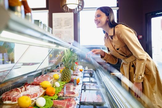 Happy young woman choosing meat from glass cabinet in grocery store