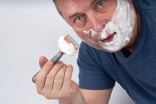 A man's face in shaving foam shaves with a safety razor