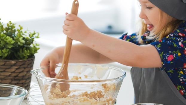 Cute little girl mixing the dough for cookies with wooden spoon in glass bowl