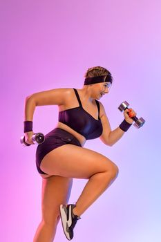Body positivity concept. Size plus model woman jumping with dumbbells. Fat girl plump girl trying to lose weight on pink background.