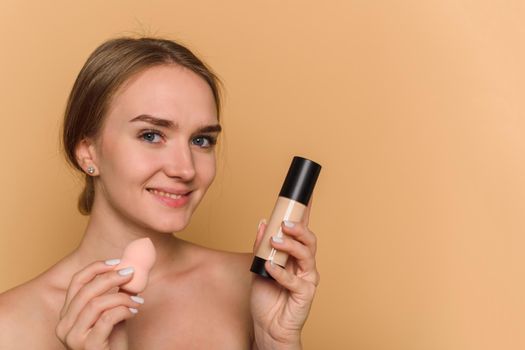 A beautiful young woman applies make-up in a beige background. Smiling lady holding sponge and foundation.