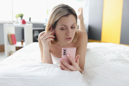 Woman lying in bed with mobile phone in her hands and inserting wireless headphones
