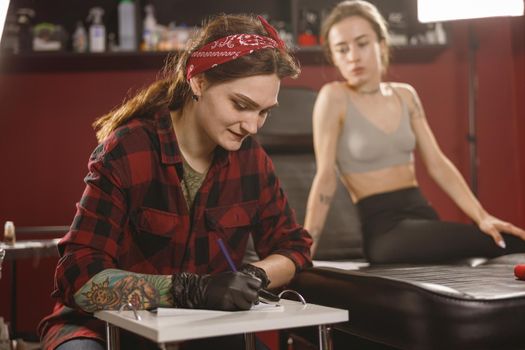Young lady in top sitting in tattoo salon and watching tattoo artist drawing