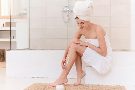 A woman applies moisturizer to her legs after a bath. A woman in a bath towel sits in a modern bathroom. Home spa treatments