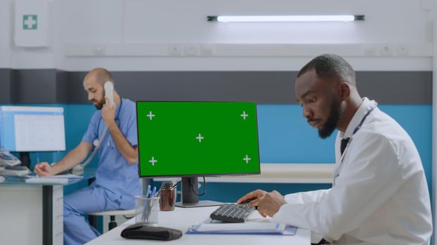 African american doctor sitting at desk in hospital office working at healthcare treatment typing medical expertise on mock up green screen chroma key computer with isolated display. Medicine concept