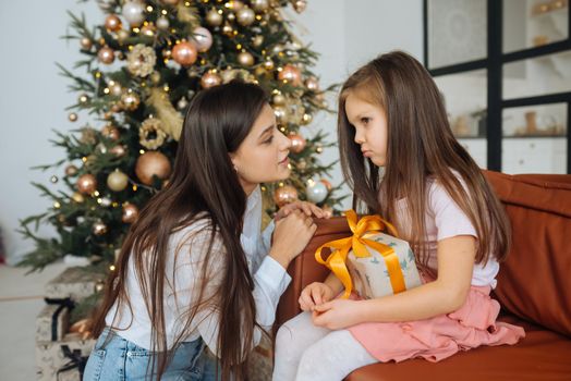 Young mother calming down her sad infant daughter sitting on the Christmas tree background