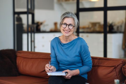 Happy senior woman in glasses writing to notebook at home