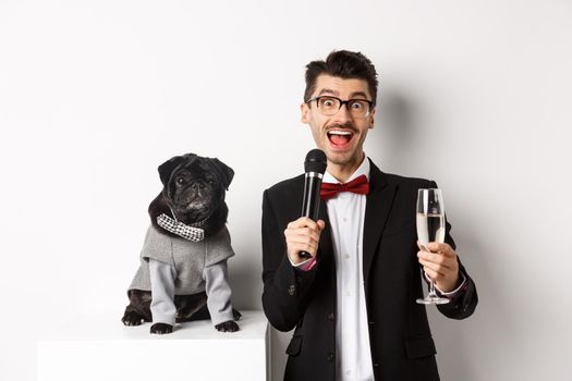 Happy young man giving a speech and raising glass champagne, standing with cute black dog in party costume over white background