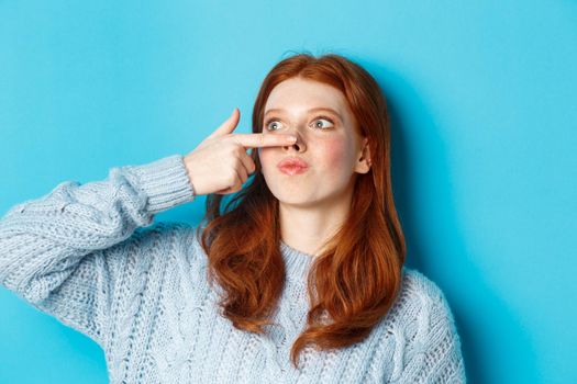 Close-up of silly redhead girl showing piggy nose, making funny grimaces and standing against blue background