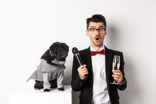 Male entertainer raising glass of champagne, giving microphone to cute black dog, standing over white background