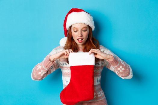 Beautiful redhead girl in santa hat open Christmas stocking and looking surprised, receiving xmas gift, standing over blue background