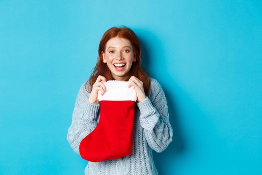 Winter holidays and gifts concept. Happy teenage redhead girl receiving xmas gift, open christmas stocking and smiling amazed, standing over blue background