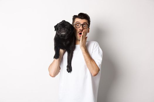 Image of shocked dog owner staring at camera and gasping impressed, cute black pug sitting on his shoulder and staring at you, white background