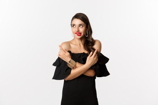 Fashion and beauty. Beautiful glamour woman in black dress, making choice, biting lip from temptation and pointing sideways, standing over white background