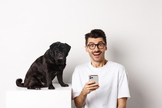 Cheerful young man hipster staring at camera, sitting with cute black pug dog and using mobile phone, standing over white background