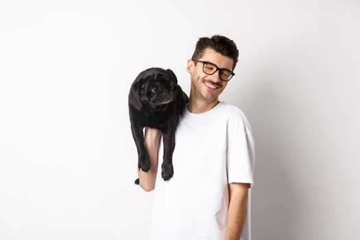 Happy man in love with dog, holding puppy on shoulder and smiling, standing over white background