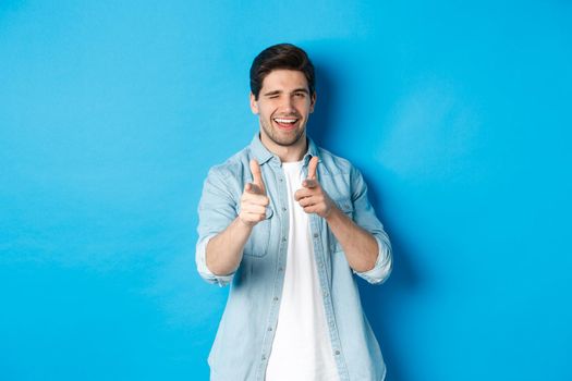 Cheeky handsome guy pointing fingers at you, winking flirty, standing in casual outfit against blue background