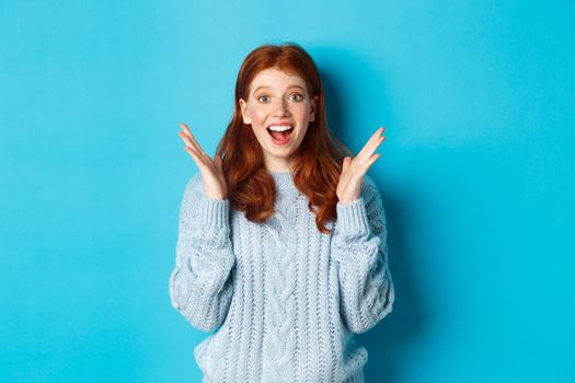 Surprised and happy redhead girl clap hands and staring at camera, smiling amazed, standing against blue background
