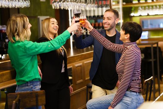 Merry multiethnic adult group of friends in casual clothes smiling and clinking glasses of alcohol drinks while resting near counter in weekend in bar
