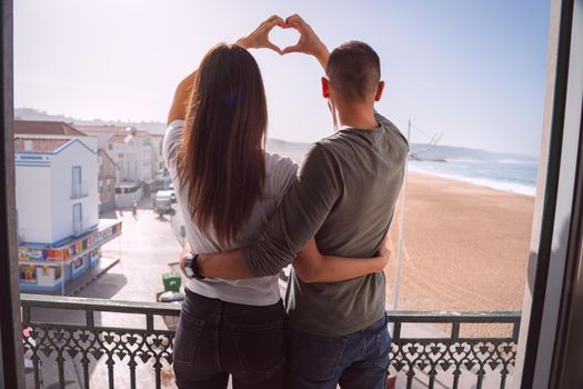 Young couple standing on the balcony and making heart shape with their hands. Couple in love hugging and spending valentine's day together