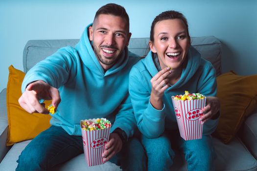 Young happy couple with popcorn sitting on sofa and watching funny comedy movie. Couple enjoying time together at home