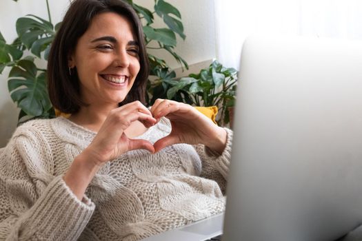 Happy young caucasian woman making heart shape with hands during online video call using laptop. Copy space.