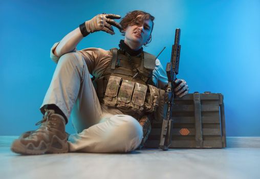 a male soldier in camouflage is sitting by an ammunition crate on the floor with a weapon