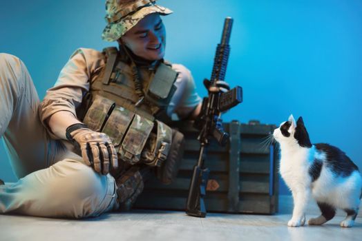 a male soldier in camouflage is sitting on the floor by a box of ammunition with weapons next to a small cat