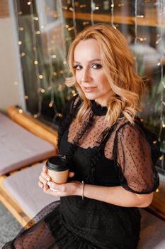 Young charming blonde with a cute smile and makeup while relaxing in a cafe. She is holding a cup of coffee in her hands. She is dressed in a black dress with transparent sleeves.