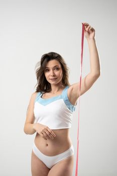 Confused brunette woman with slim body in white underwear on a diet looking in a camera and showing a pink centimeter tape. Healthy lifestyles concept. Sport and diet.
