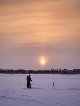 A young man fishes from a hole in the ice at dawn. winter fishing.