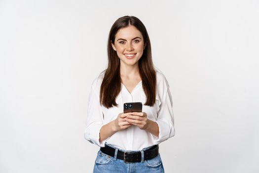 Young woman with smartphone, smiling and looking at camera, using mobile phone app, cellular technology and online shopping concept, white background