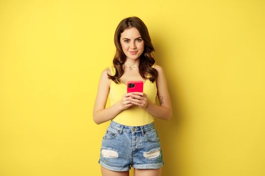 Cellular technology and online shopping concept. Young stylish woman using smartphone, mobile phone application, standing over yellow background