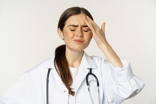 Portrait of distressed, frustrated doctor, holding hands on forehead and grimacing from headache, painful migraine, standing over white background