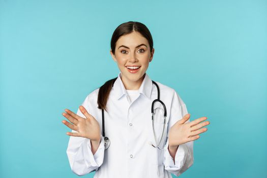 Happy and excited smiling young woman looking with excitement and surprised, standing in white lab coat and stethoscope, blue background