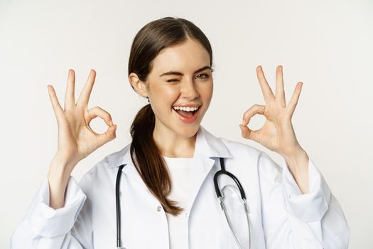 Happy beautiful woman doctor, showing okay, ok excellent sign, smiling amazed, approve smth, praise, standing in white lab coat over white background