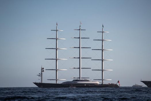 A huge sail yacht of conceptual design with three masts stands in the bay near Monaco at sunny day, the sails are lowered, the yacht spins around its anchor, left side of the sailboat, clear weather