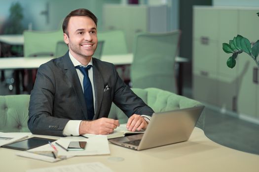 Handsome man in business suit working on laptop, job in progress. Cheer smiling freelancer making notes in scheduler sitting in bright coworking space