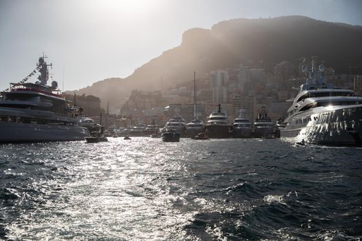 A lot of huge yachts are in port Hercule of Monaco at sunset, mountain is on background, magic backlight is on glossy boards of the motor boats, megayachts are moored in marina, sun reflections
