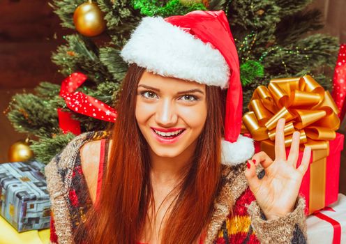 New Year Woman with Long Hair in a Red Hat Gazes to the Camera and Shows Hand Ok Sign. Green Decorated New Year Tree with lots of Gift Boxes Background. Close-up