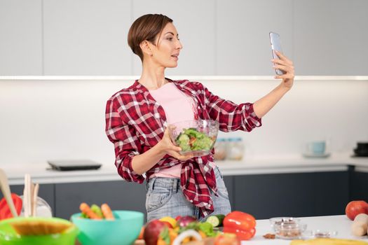 Pretty housewife taking selfie or making a video call using her smartphone while cooking fresh salad wearing a plaid shirt with a bob hair style. Healthy food leaving - vegan concept