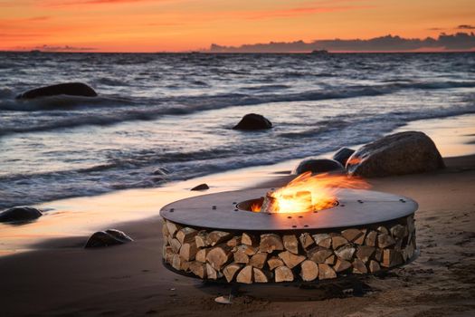 The beautiful metal round a fireplace stands on the sandy coast with a tidal wave at sunset, stones, sand, waves, the landscape, tranquillity, the sun falls, firewood, nobody