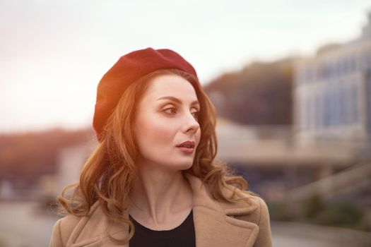 Charming pretty french woman in an autumn beige coat and red beret standing outdoors with urban city background. Tinted photo