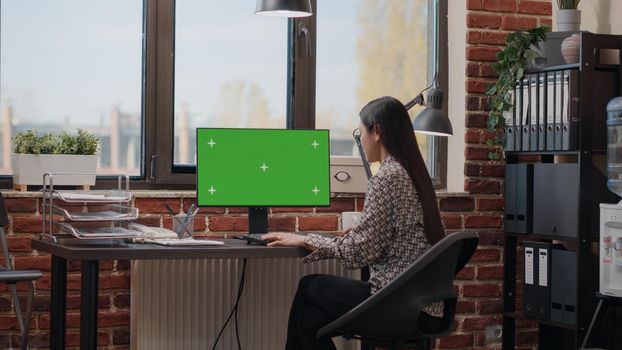 Asian woman looking at horizontal green screen on computer in business office. Company employee using monitor with chroma key and isolated mockup template to plan marketing strategy.