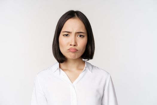 Sad and gloomy young asian woman grimacing, frowning upset, making pouting face, white background