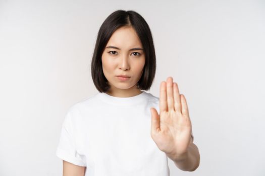 Image of asian girl showing stop, prohibit smth, extend one arm to show forbidding, taboo gesture, standing in tshirt over white background