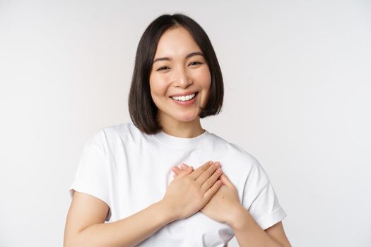 Close up of smiling korean woman holding hands on heart, care and love concept, feel affection, tenderness or heartwarming feeling, standing over white background