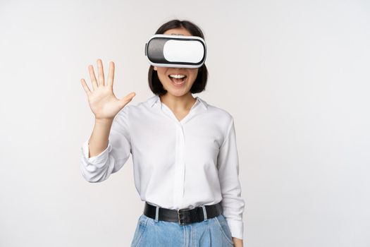 Smiling asian woman waving at you, wearing virtual reality glasses, vr headset, chatting in meta universe, standing over white background